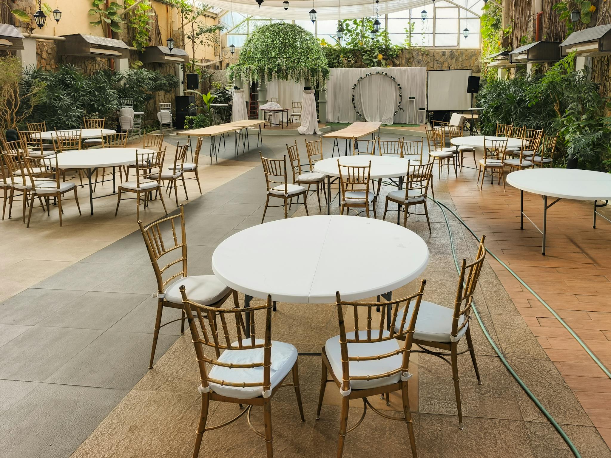 Empty elegant indoor venue setup with round tables and chairs, greenery, and soft lighting.